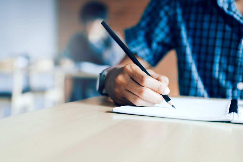 Close up of teenager student hand writing on paper exam with a pencil in classroom.